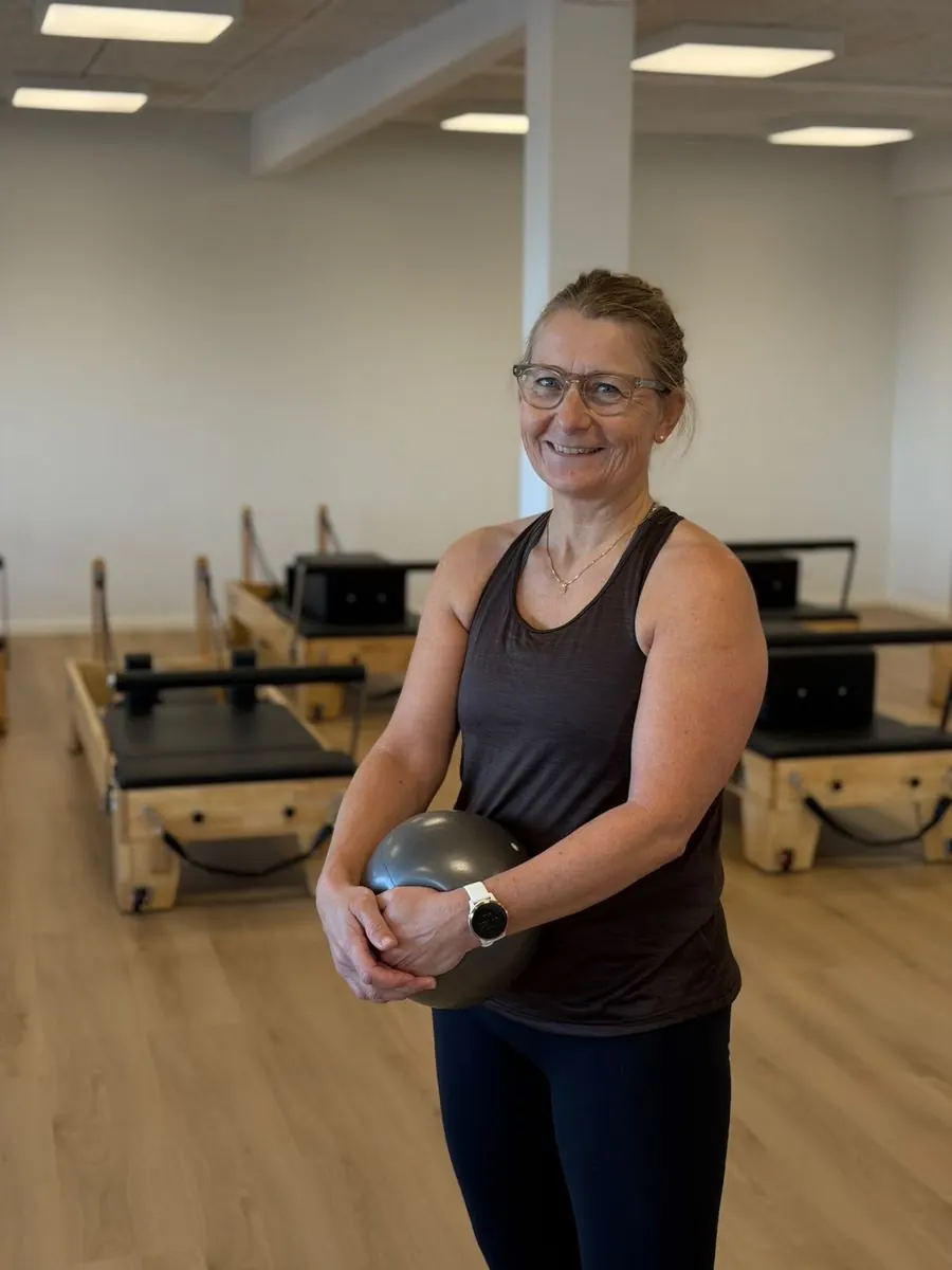 Jane Clausen smiler med en pilatesbold i hænderne foran studiets Reformer-maskiner